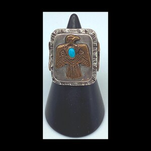 Op de afbeelding: Een zilveren ring met een vierkant ontwerp met een turquoise steen en een gouden adelaar met ingewikkelde details.
