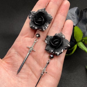 Puede incluir: Un par de pendientes de rosa negra con dijes de espada plateada y detalles de cristal negro.