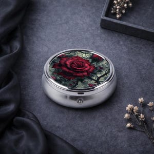 Könnte beinhalten: Eine runde, silberne Pillendose mit einem detaillierten, dunkelroten Rosenmuster auf dem Deckel. Die Rose ist vor einem grau-grünen Hintergrund mit filigranen, rankenartigen Details platziert. Die Dose hat einen kleinen, silbernen Knopf auf der Vorderseite.