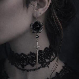 Puede incluir: Pendiente plateado con una rosa negra y un colgante de espada. El pendiente tiene un diseño gótico. La rosa tiene pétalos texturizados, y la espada tiene detalles de mango. Una cuenta negra conecta la rosa con la espada. Joya única.