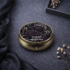 Könnte beinhalten: Eine runde Schmuckdose im antiken Stil mit goldener Oberfläche. Der Deckel zeigt ein Design aus lila Rosen und die Worte "POISONS AND ANTIDOTES" in einer gotischen Schrift. Die Dose befindet sich vor einem dunkelgrauen Hintergrund.