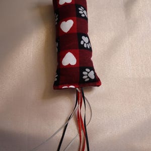 May include: Red and black plaid cat toy with white paw prints and hearts. The toy has a long tail with red, white, and black ribbons.