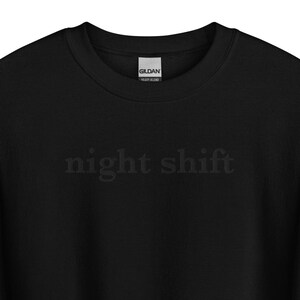 May include: Black crew neck sweatshirt with the text "night shift" in a tonal, slightly raised font across the chest. The sweatshirt has a ribbed collar, cuffs, and hem. The Gildan brand label is visible at the back of the neck.