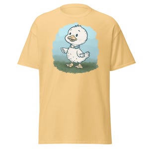 May include: A light yellow t-shirt featuring a cartoon duck illustration. The duck is white with a blue collar and stands on green grass against a light blue background. The shirt is a solid color.