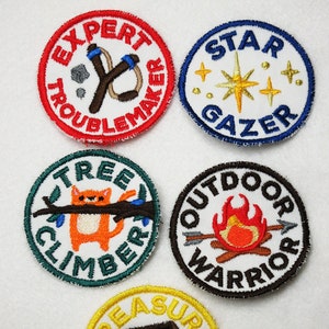 以下が含まれることがあります： 白い背景に異なるデザインが施された、カラフルな刺繍パッチが5つ。パッチには、「エキスパートトラブルメーカー」とスリングショット、「スターゲイザー」と星、「ツリークライマー」と猫、「アウトドアウォリアー」と焚き火、「トレジャーハンター」と宝箱が描かれている。