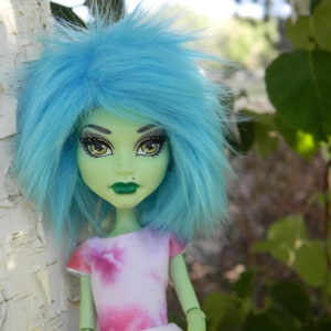 Puede incluir: Una muñeca de piel verde con cabello azul y ojos verdes. Lleva una camisa de color rosa y blanco con estampado tie-dye.