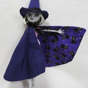 May include: A purple and black doll wearing a witch hat and a cape with a black cat print. The doll has black eyes and white skin.