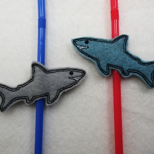 Op de afbeelding: Twee vilt haaienvinnen dranktoppers, een blauwe en een grijze, bevestigd aan blauwe en rode rietjes.