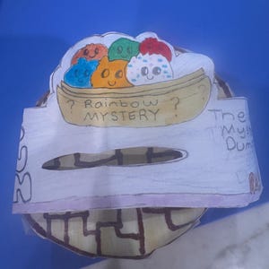 May include: A handmade paper craft featuring a boat filled with colorful, smiling dumplings. The text "? Rainbow Mystery ?" is written below the boat. The craft has a circular base with a maze-like design.