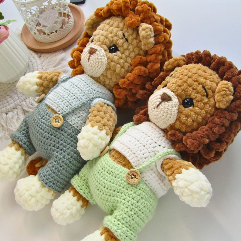 May include: Three handmade crochet lion plush toys. The lions have brown manes, cream-colored faces, and black eyes. Two lions wear overalls in light green and gray, with wooden button closures. The plush toys are soft and cuddly.