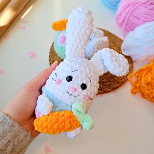 Puede incluir: Un conejito de crochet blanco sosteniendo una zanahoria naranja brillante. El conejito tiene ojos negros y una nariz rosa. La zanahoria tiene un tallo verde.