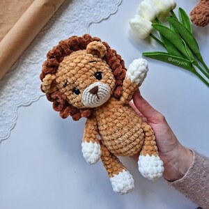 May include: A handmade crochet lion plush toy in shades of brown and tan. The lion has a fluffy mane, a cream-colored muzzle, and white accents on its paws. The toy is soft and cuddly, perfect for a child's room or nursery.