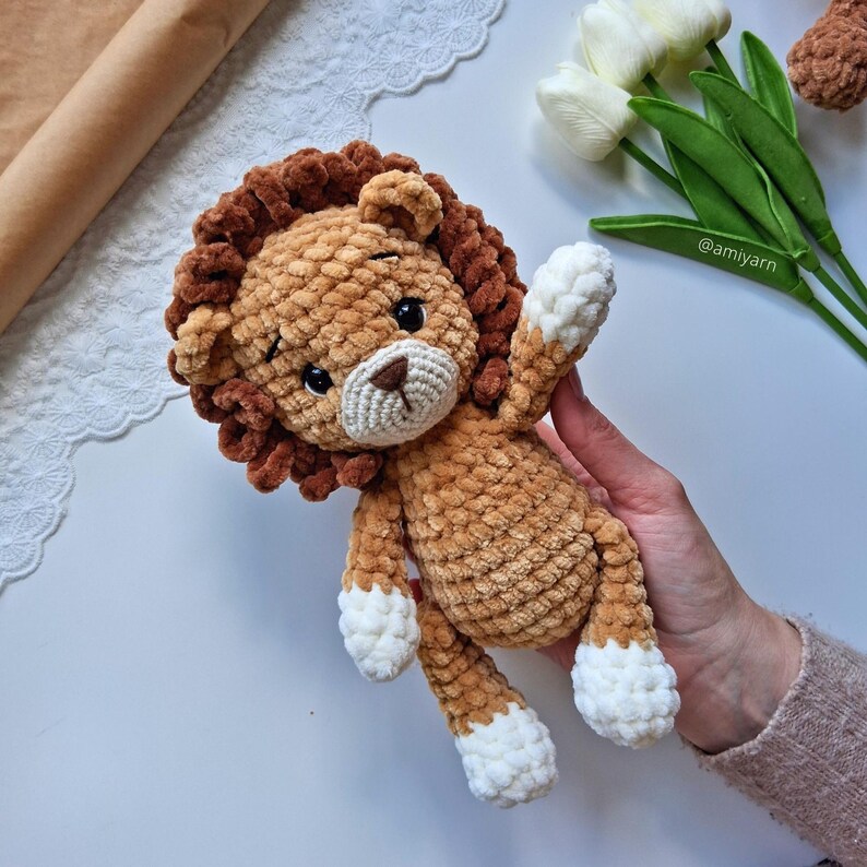 May include: A handmade crochet lion plush toy in shades of brown and tan. The lion has a fluffy mane, a cream-colored muzzle, and white accents on its paws. The toy is soft and cuddly, perfect for a child's room or nursery.