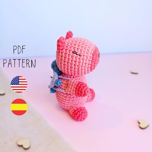 May include: Pink crocheted amigurumi toy with a blue and purple backpack. The toy has closed eyes and a smiling mouth. The toy is sitting on a pink surface. PDF PATTERN is written in black text on a white background.