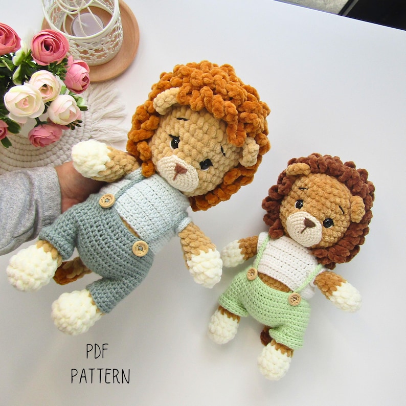 May include: Two handmade crochet lion dolls. One lion wears a light blue outfit with wooden buttons, the other wears a green outfit. Both have brown manes, tan faces, and white paws. The text "PDF PATTERN" is visible.