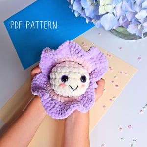 Puede incluir: Una flor de ganchillo hecha a mano con una cara sonriente. La flor tiene un aspecto esponjoso y texturizado con tonos de morado, rosa y blanco. El texto "PDF PATTERN" es visible en el fondo. La flor se sostiene en las manos.