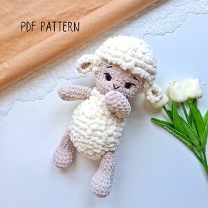 Puede incluir: Un juguete de peluche de cordero blanco y beige tejido a crochet con una textura esponjosa. El cordero está acostado de lado con la cabeza inclinada. El cordero tiene ojos negros y una nariz rosa.