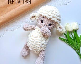 May include: A crocheted white and beige lamb plush toy with a fluffy texture. The lamb is lying on its side with its head tilted to the side. The lamb has black eyes and a pink nose.