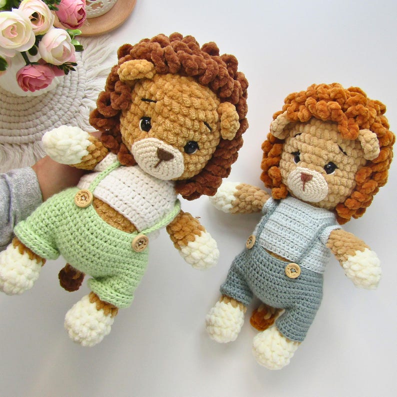 May include: Two handmade crochet lion dolls. One lion wears green overalls with wooden buttons, the other wears blue overalls. Both have brown manes, tan faces, and white and tan paws.