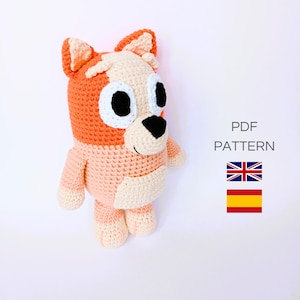 Puede incluir: Patrón de crochet para un perro de peluche. El perro es naranja, rosa y blanco con ojos negros y una nariz negra. El patrón está disponible en inglés y español.