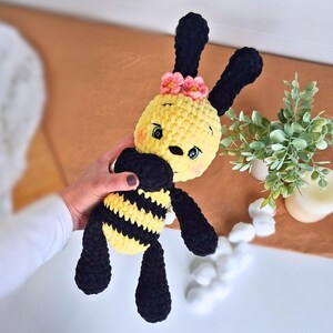 Puede incluir: Un peluche de abeja hecho a mano con ganchillo. La abeja tiene la cabeza y el cuerpo amarillos con rayas negras, antenas negras y patas negras. Tiene una flor rosa en la cabeza.