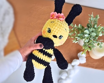 May include: A handmade crochet bee plush toy. The bee has a yellow head and body with black stripes, black antennae, and black legs. It has a pink flower on its head and is being held by a person.