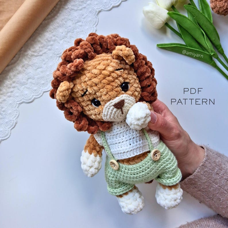 May include: A handmade crochet lion plushie with a brown mane, white muzzle, and green overalls. The lion is holding a white ball and has the text "PDF PATTERN" in the image. The plushie is a soft, cuddly toy.