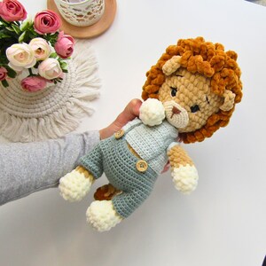 May include: A handmade crochet lion doll with a fluffy orange mane, beige face, and light blue overalls. The doll has button details and cream-colored paws. A soft toy for children.