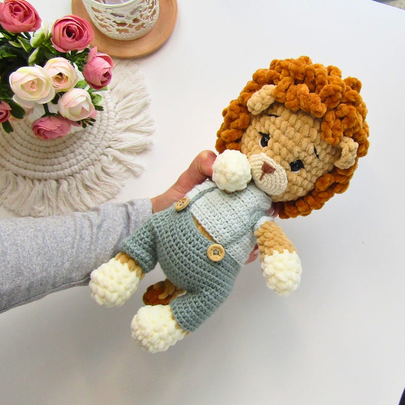 May include: A handmade crochet lion doll with a fluffy orange mane, beige face, and light blue overalls. The doll has button details and cream-colored paws. A soft toy for children.
