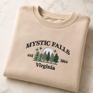 Könnte beinhalten: Ein beigefarbenes Sweatshirt mit Rundhalsausschnitt und schwarzer Stickerei mit der Aufschrift Mystic Falls Virginia sowie der Abkürzung est 1864. Das Design zeigt einen Wald aus grünen Tannen, einen weißen Vollmond und kleine schwarze Sterne.