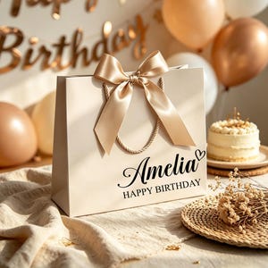 Może przedstawiać: Kremowa torebka prezentowa z dużą, pasującą satynową kokardą. Na torebce nadrukowano imię Amelia oraz napis HAPPY BIRTHDAY czcionką pisankową i bezszeryfową, a także małą ikonę serca.