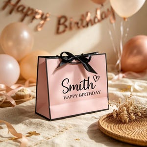 Może przedstawiać: Jasnor&oacute;żowa torebka prezentowa z czarnym wykończeniem i czarną satynową kokardą. Z przodu znajduje się czarny napis "Smith" nad "HAPPY BIRTHDAY" oraz mała ikona serca. Torebka posiada solidne dno i czarne uchwyty ze sznurka.