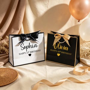 Może przedstawiać: Dwie prostokątne torebki prezentowe z uchwytami ze wstążki. Biała torebka z czarną kokardą, imieniem Sophia i napisem Happy 21st Birthday z sercem. Czarna torebka ze złotą kokardą, imieniem Olivia i napisem Happy 45th Birthday z sercem.