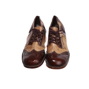 May include: A pair of two-tone oxford shoes featuring a dark brown wingtip toe cap with decorative perforations, a tan mid-section, and dark brown lace-up panels. The shoes have dark brown laces and brass-colored eyelets for a classic look.