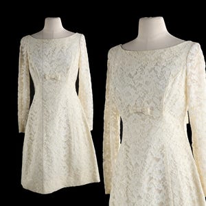 May include: An off-white, knee-length dress featuring long sleeves, a boat neckline, and an all-over floral lace overlay. The garment is fitted at the waist and includes a small, centered decorative bow detail on the front bodice.