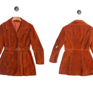 May include: A burnt orange suede jacket featuring a collared neckline, long sleeves, and a front button closure. The jacket includes a matching belt with a gold-tone buckle, two front patch pockets, and visible topstitching throughout the garment.