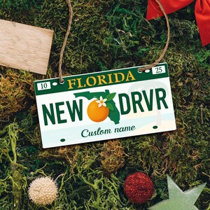 May include: A Florida-themed license plate ornament with the text "NEW DRVR" and a graphic of an orange and the state outline. The ornament has a green and white color scheme, with the word "FLORIDA" at the top. A custom name can be added.