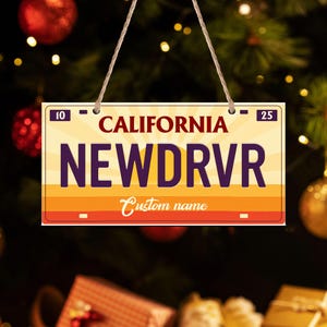 May include: A California license plate-shaped ornament with the text "NE WDRVR" in purple, and "Custom name" in cursive. The background features a sunset design with orange and yellow stripes. The ornament is hanging by a string.