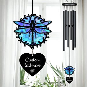 May include: A decorative wind chime featuring a dragonfly design with blue and purple stained glass. A black heart-shaped tag with the text "Custom text here" hangs below. The wind chime has black metal tubes.