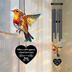 May include: A stained-glass style robin bird suncatcher with a heart-shaped memorial. The heart reads "When a robin appears, a loved one is near" and "Mom 1955-2025". A black wind chime hangs in the background.