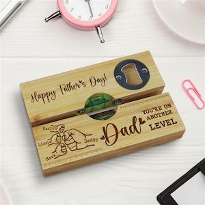 May include: A wooden bottle opener with the words "Happy Father's Day!" and "Dad" engraved on it. It features a bottle opener and a decorative green and clear glass piece. The names Katlyn, Lizzie, and Mark are also engraved.