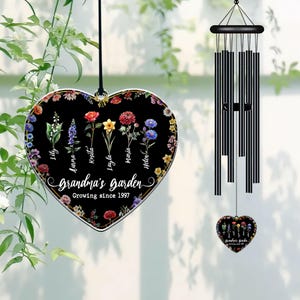 May include: A black heart-shaped wind chime with a floral design and the text "Grandma's Garden Growing since 1997". The chime has a silver border and features the names of different flowers, including Lily, Moira, Krista, Layla, Maria, and Helen.