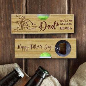 Pu&ograve; includere: Due apribottiglie in legno con livelle a bolla. Uno dice "Pap&agrave;" con nomi e "Sei a un altro livello". L'altro dice "Buona festa del pap&agrave;!" e ha un apribottiglie. Lo sfondo &egrave; una superficie di legno.