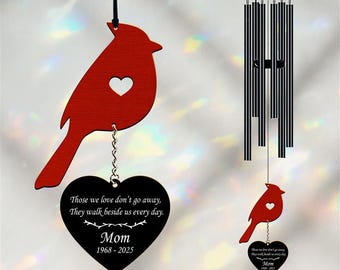 May include: A red cardinal-shaped ornament with a heart cutout, hanging from a black string. A black heart-shaped pendant with the text "Those we love don't go away, They walk beside us every day. Mom 1968-2025" hangs below. A wind chime is also visible.