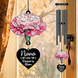 May include: A pink and white carnation wind chime with the names "Eva", "Taylor", "Simon", "Sophi", "John", "Katie", and "Ashley" on the petals. The chime has a black heart-shaped charm with the text "Nana Forever in Bloom".