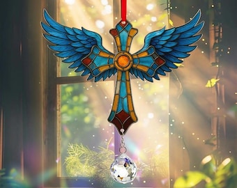 May include: A stained glass cross with blue angel wings, a central orange circle, and a clear crystal pendant. The cross is suspended by a red ribbon, creating a decorative suncatcher for a window.