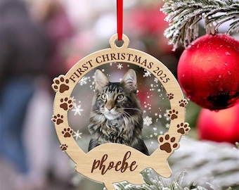 May include: A wooden Christmas ornament featuring a cat named Phoebe. The ornament has a circular design with the text "First Christmas 2025" and paw prints. A red ribbon is attached for hanging. The cat has brown and black fur.