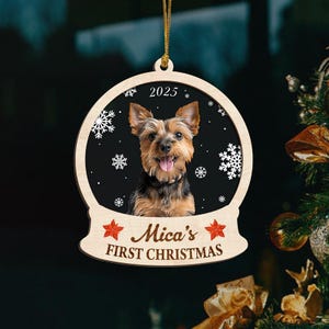 May include: A wooden Christmas ornament shaped like a snow globe, featuring a Yorkshire Terrier dog. The ornament has the text "2025" at the top and "Mica's FIRST CHRISTMAS" at the bottom. Snowflakes and red star accents decorate the black background.