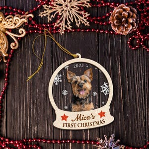 May include: Wooden Christmas ornament shaped like a snow globe, featuring a photo of a dog with the text "2025" and "Mica's FIRST CHRISTMAS". The ornament is decorated with snowflakes and a gold string for hanging.