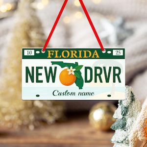 May include: A Florida license plate ornament with a red ribbon for hanging. The plate features the state name, a green outline of Florida, an orange, and the text "NEW DRVR". The bottom of the plate reads "Custom name".